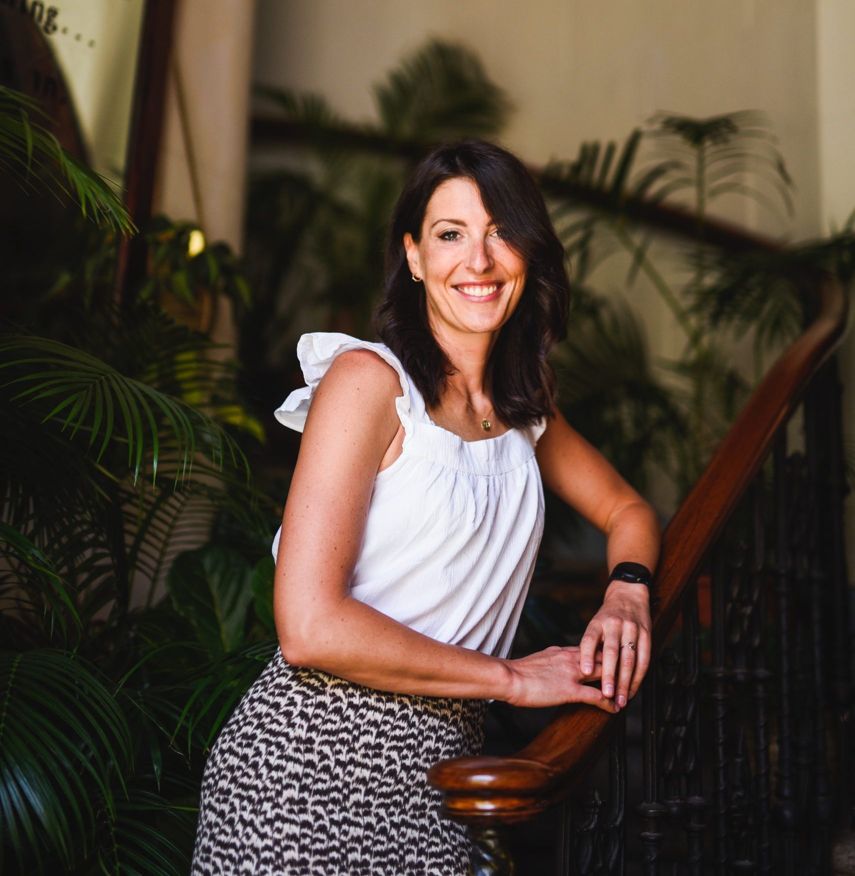 Karolina leaning on a bannister. She is a white lady with brown hair and is smiling, behind her are plants.